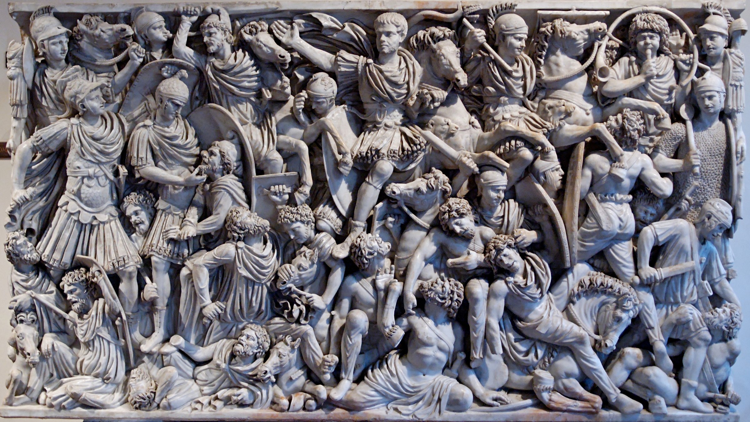 Marble relief crowded with fighting figures: Roman horsemen and soldiers in helmets and cloaks ride over bearded barbarian warriors, who fall, kneel and struggle beneath them
