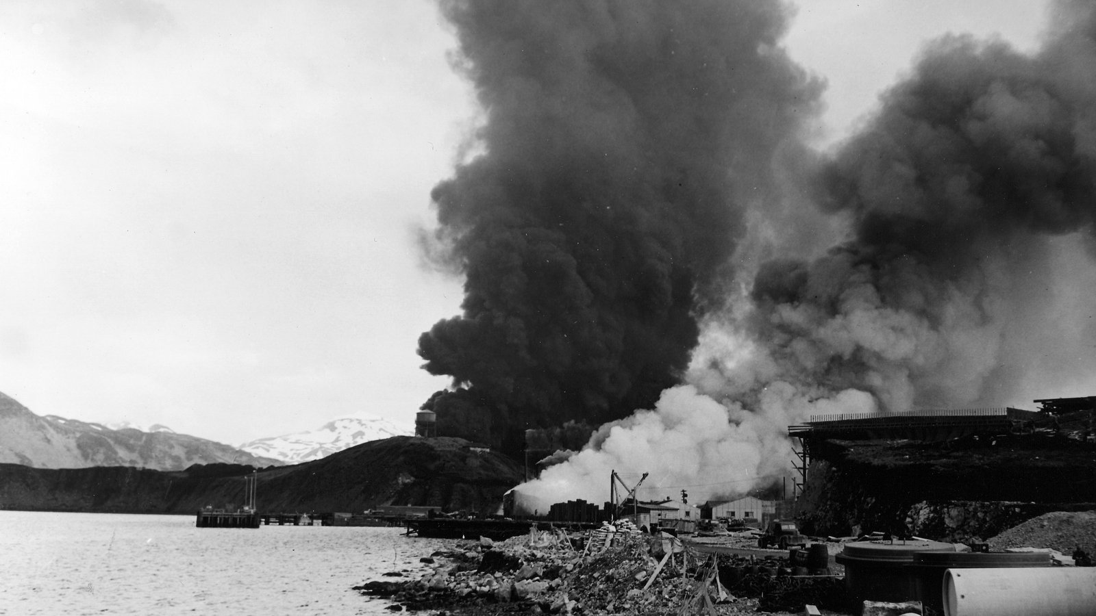 Black and white photograph of huge columns of black smoke and white steam rising from burning oil tanks and a ship beside a rocky shore, with snowy mountains behind.