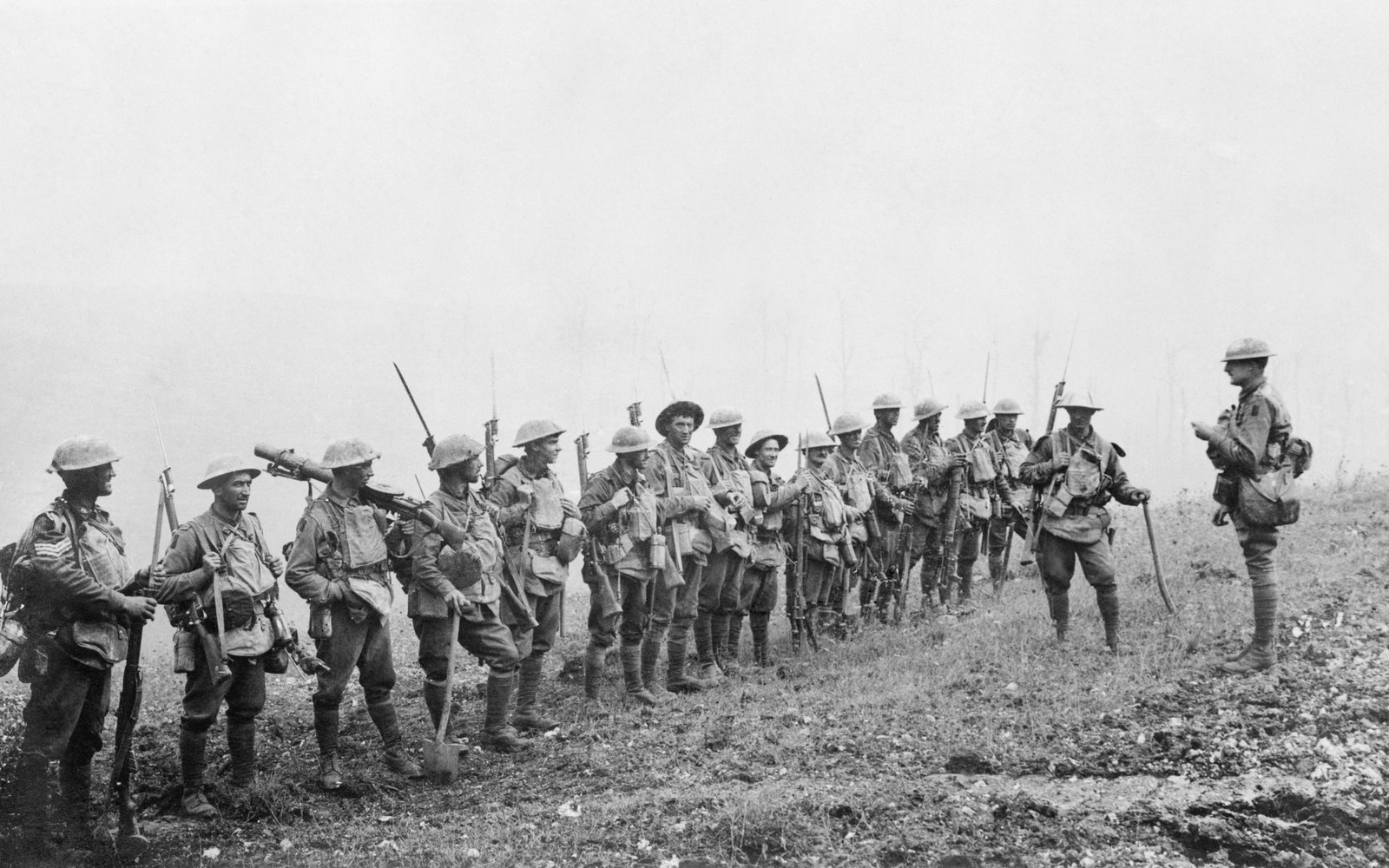 Black and white photograph of a line of Australian infantrymen in steel helmets and one slouch hat, rifles with fixed bayonets and a Lewis gun on their shoulders, standing on a grassy slope in thick white mist while an officer on the right addresses them