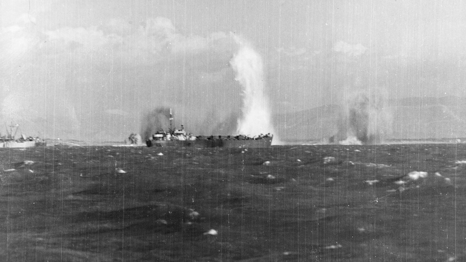 Black and white photograph from a ship at sea: a landing ship low on the water, a tall white column of spray from a bomb burst behind it, and hills on the horizon.