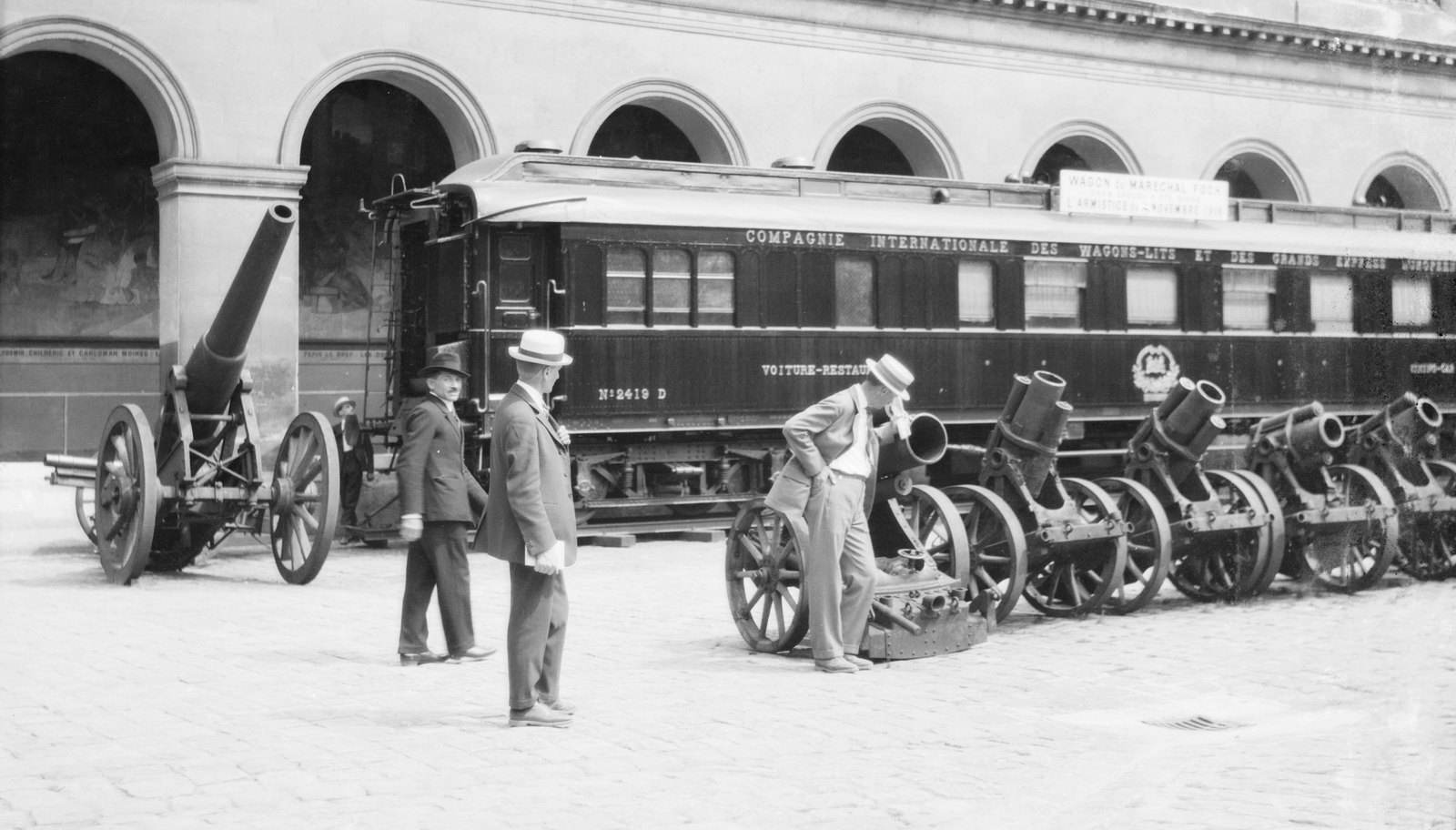 Black and white photograph of a long wooden railway dining car marked Compagnie Internationale des Wagons-Lits in a paved courtyard with arcades, with captured guns and mortars in front and men in straw hats looking at them