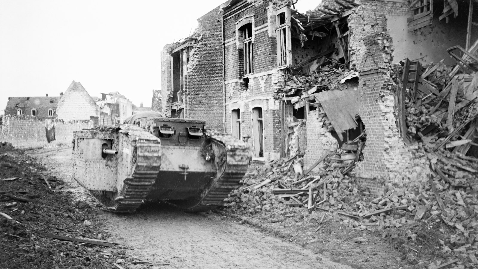 Black and white photograph of a British First World War tank rolling along a rubble-strewn street in Arras past shattered brick houses.