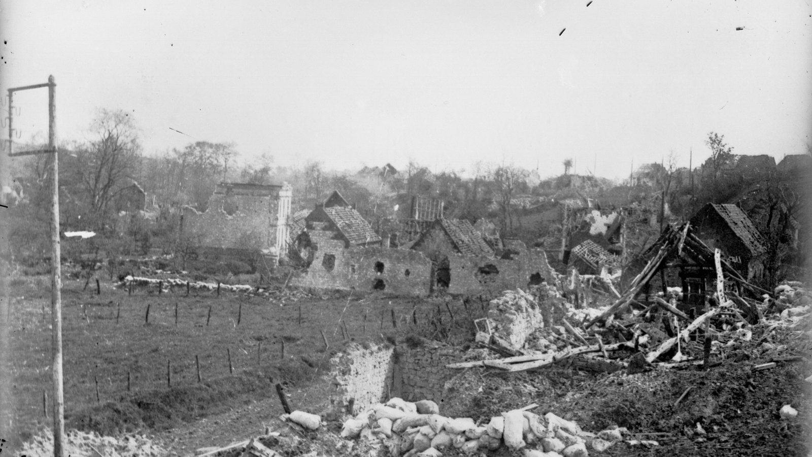 Black and white 1915 photograph of the shell-wrecked village of Carency in Artois: roofless stone houses and shattered timber beyond a field of barbed wire, with sandbags and debris in the foreground.