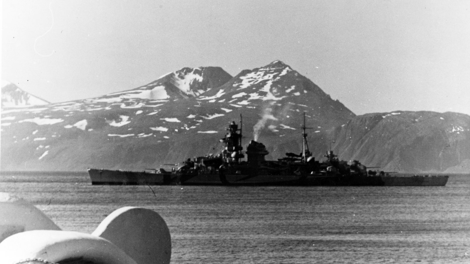 Black and white photograph of a long, camouflaged German heavy cruiser at anchor in a calm fjord, with snow-streaked mountains behind and a ship's deck fittings in the foreground.