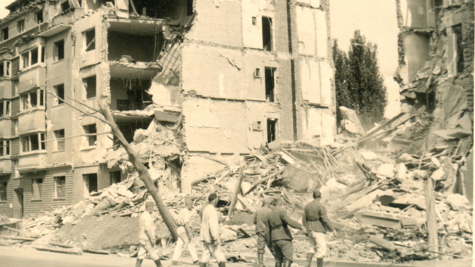 Old photograph of a five-story apartment block in Belgrade with its front torn away, a mountain of rubble and broken beams in the street, and a few soldiers and civilians walking past.