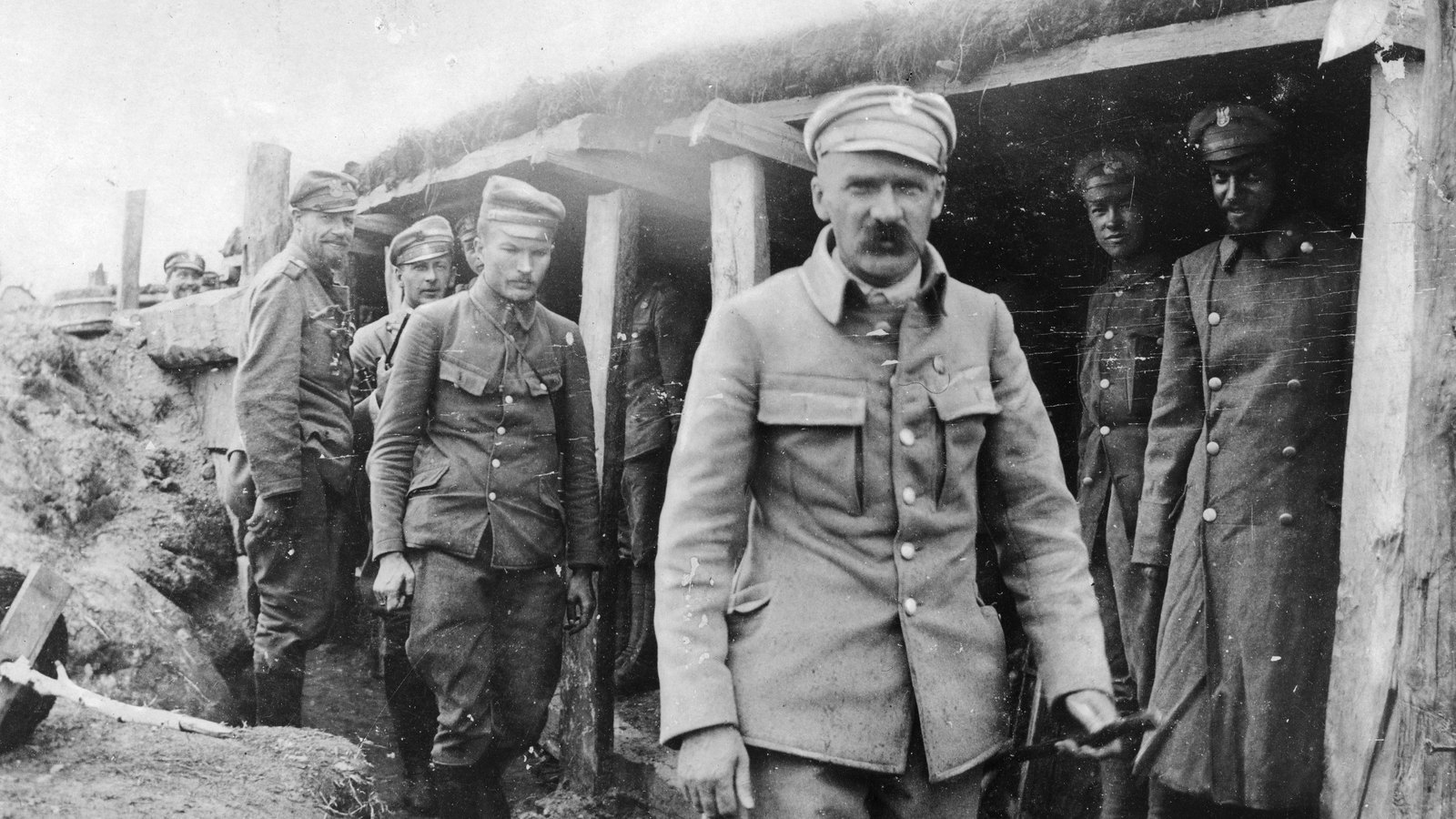 Black and white photograph of Józef Piłsudski in a peaked cap walking out of a log-roofed trench shelter, with officers of the Polish Legions behind him.