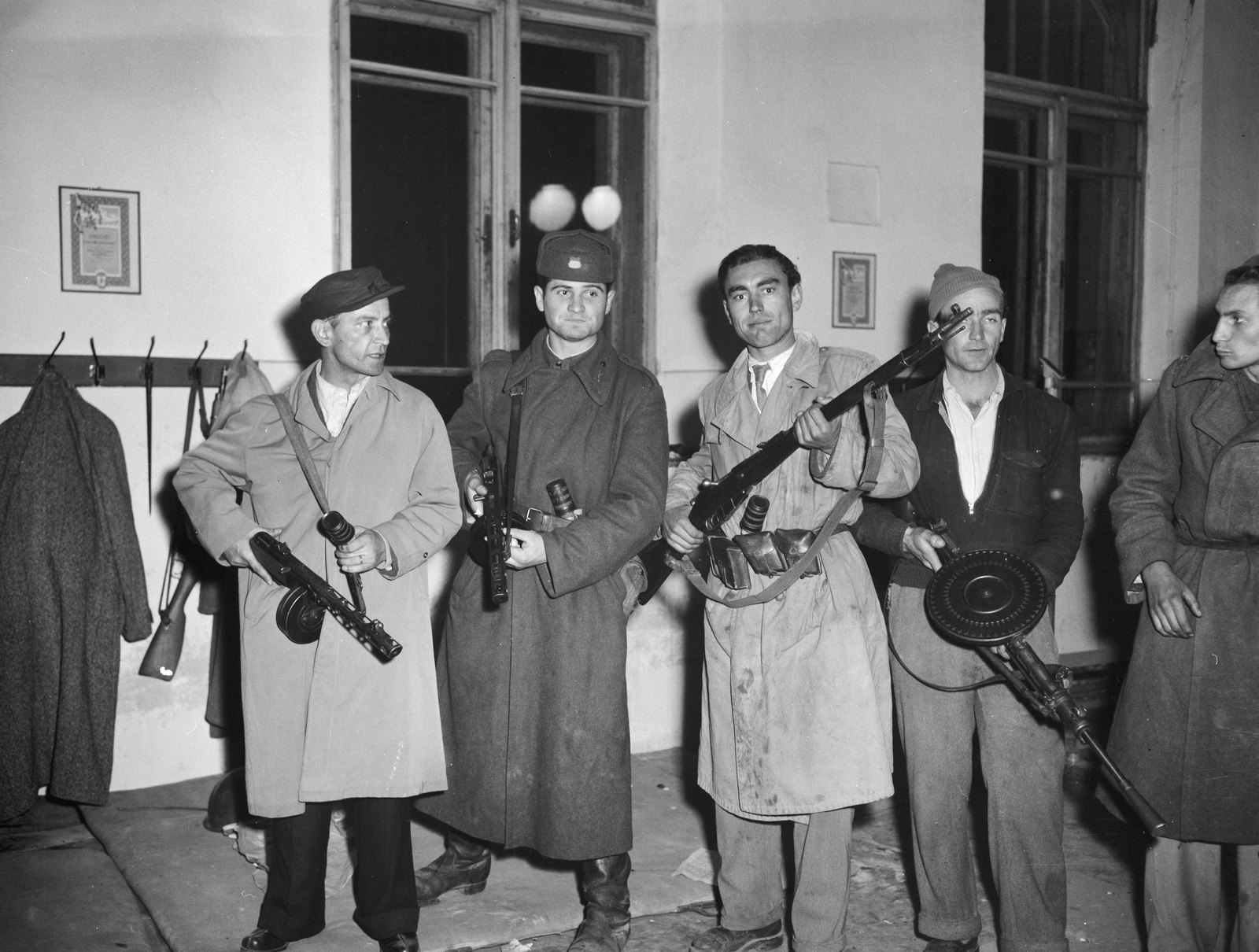 Black and white photograph of five armed men in long coats and caps holding submachine guns with drum magazines in front of a white building with tall windows and coats hanging on wall hooks