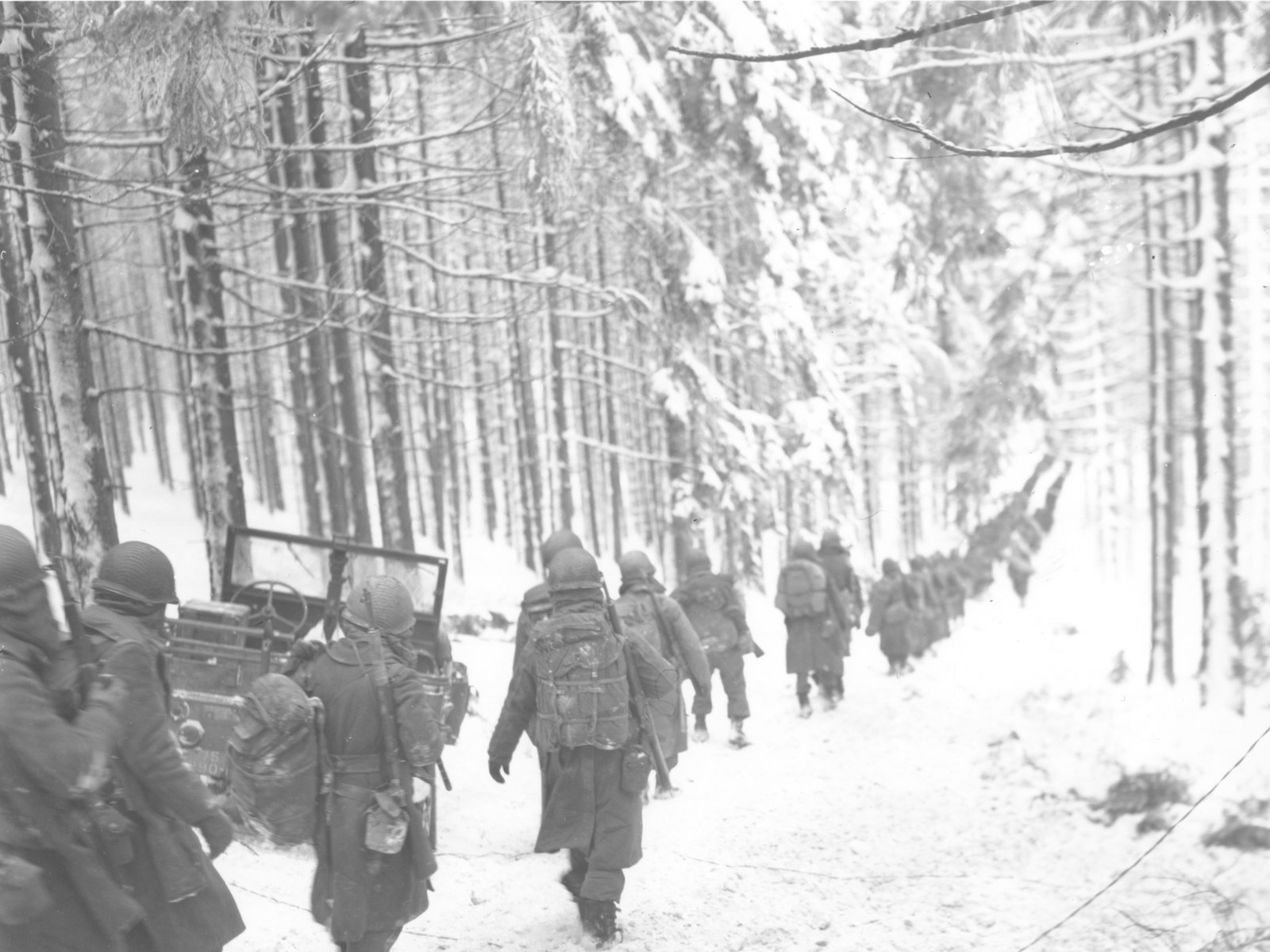 American soldiers of the 75th Infantry Division in helmets and greatcoats marching in single file along a snow-covered forest road in Belgium, January 1945