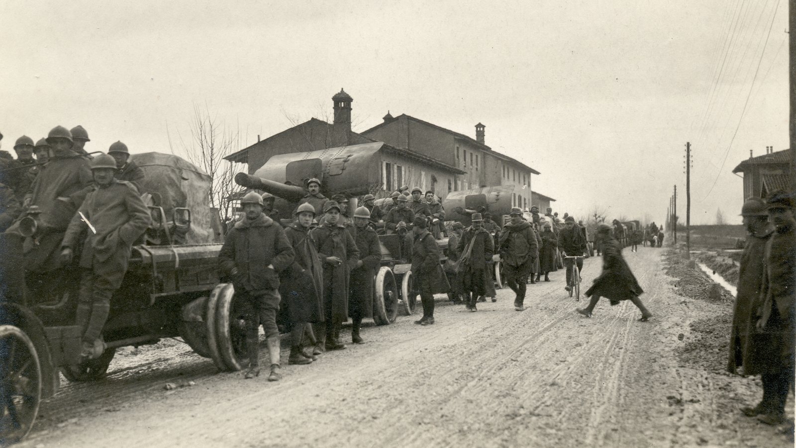 Black and white photograph of Italian soldiers in steel helmets and long coats crowding a muddy road beside trucks carrying tarpaulin-covered guns, with farm buildings and telegraph poles behind
