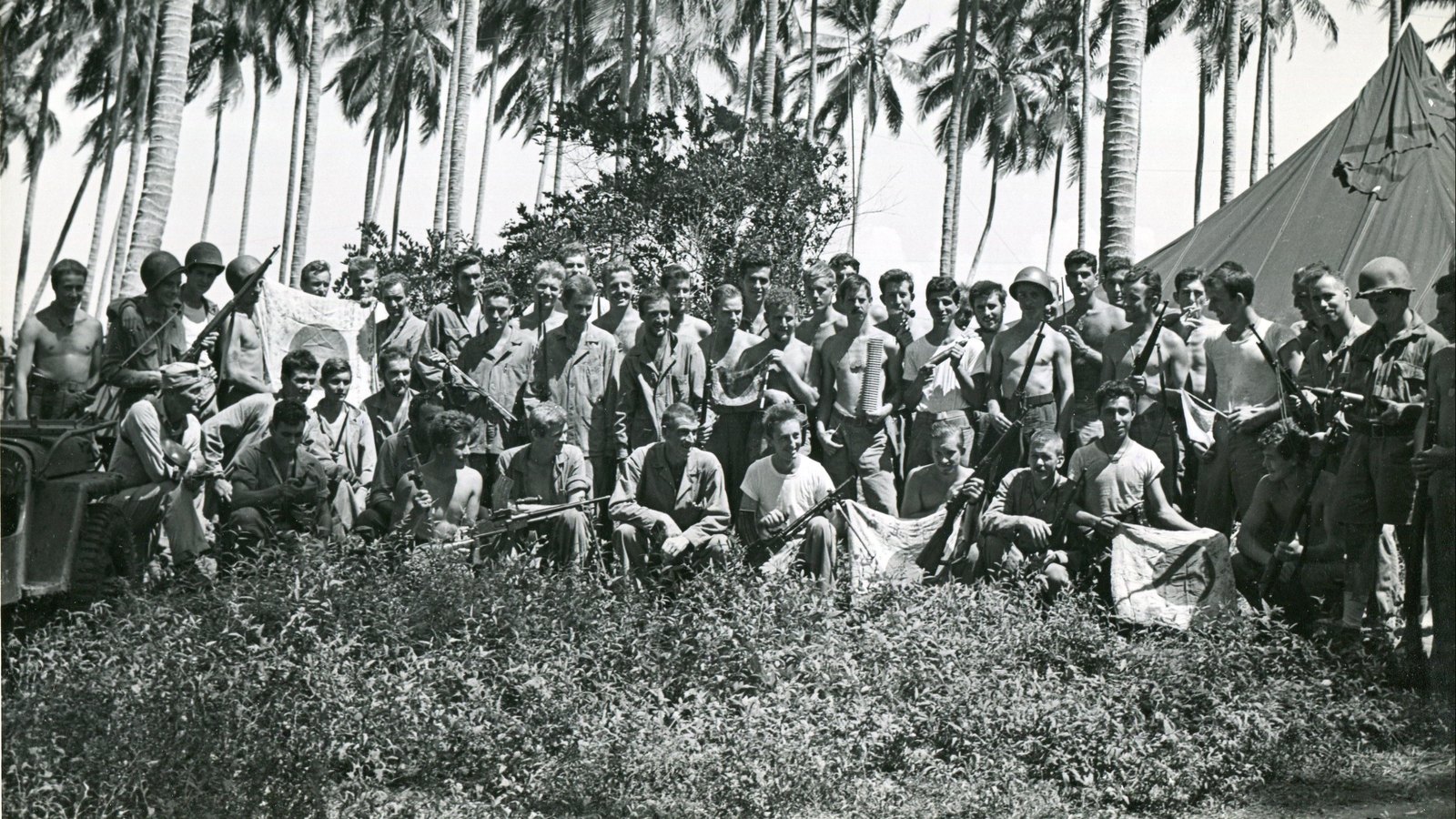 Black and white photograph of some fifty Marines, many shirtless or in helmets, posed under tall coconut palms beside a tent, holding rifles, machine guns and captured Japanese flags.