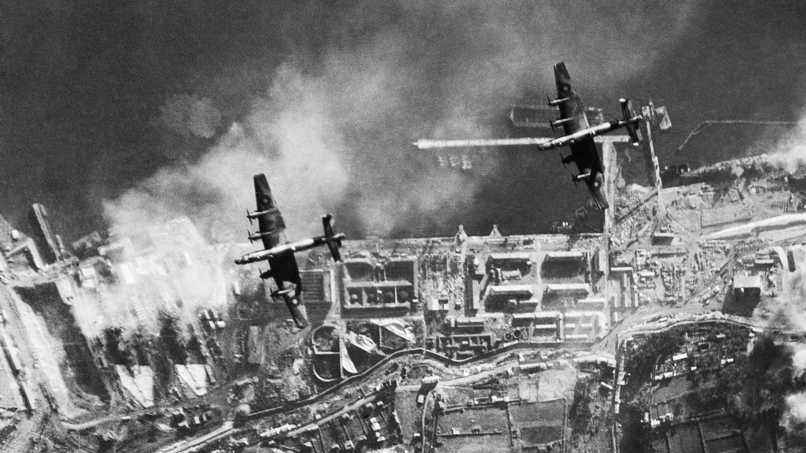 Aerial black and white photograph of two four engined bombers flying over a harbor with dry docks, a breakwater and rows of buildings, smoke drifting across the water.