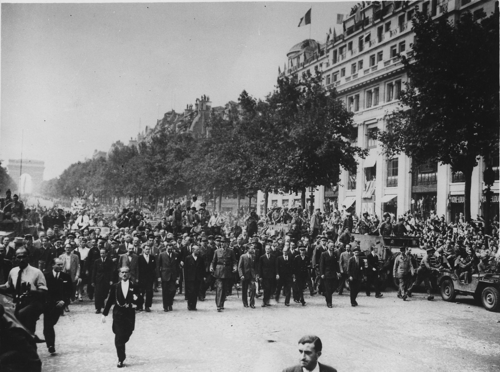 Black and white photograph of General de Gaulle in uniform walking at the head of a crowd of men in suits down the Champs-Élysées, with the Arc de Triomphe far behind and spectators on vehicles to the right