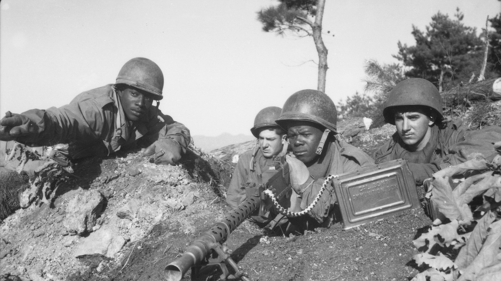 Black and white photograph of four American soldiers in steel helmets in a hilltop foxhole, one pointing ahead, behind a machine gun with an ammunition belt running from a metal box.