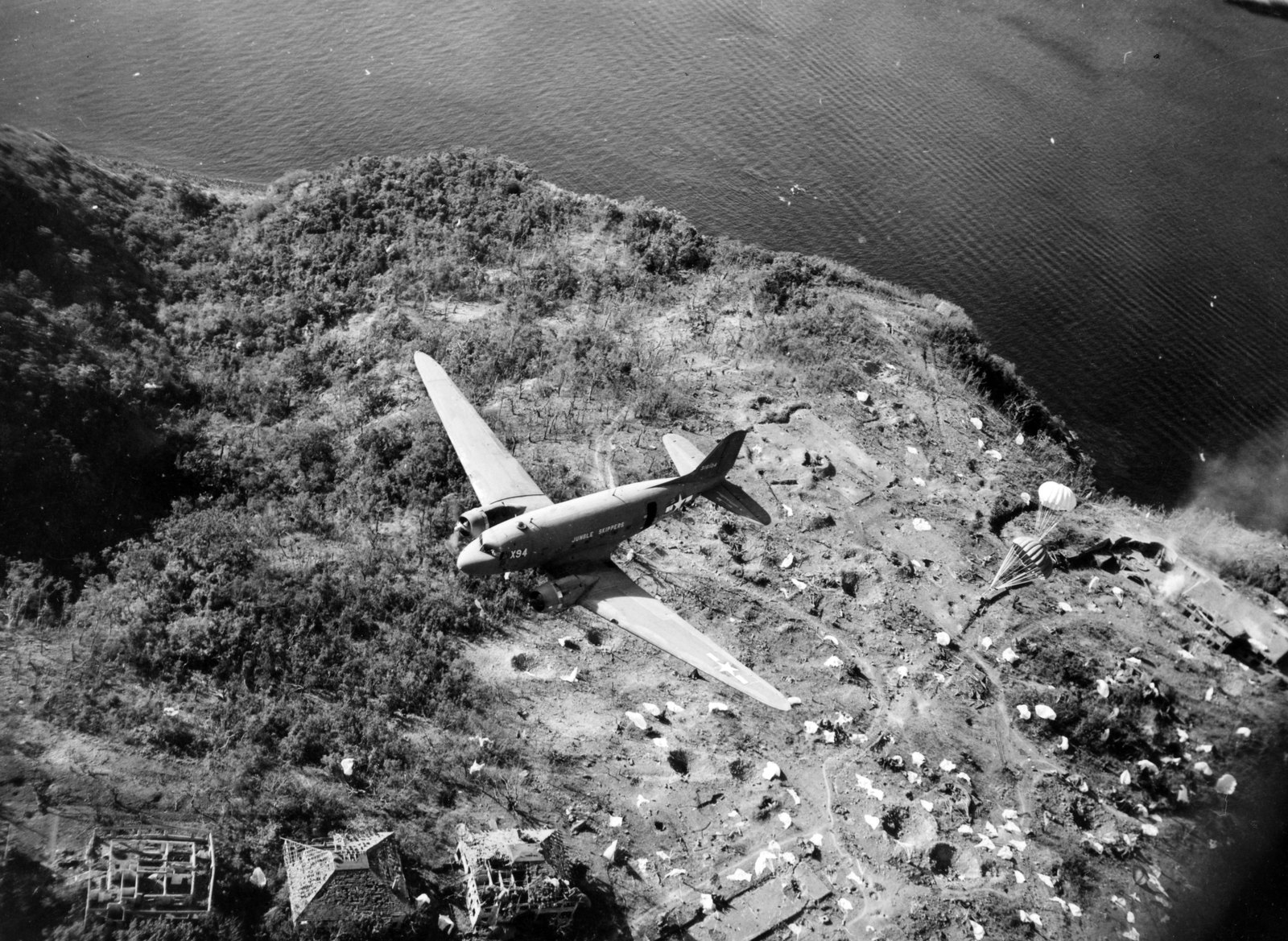 Aerial black and white photograph of a twin engine transport plane flying low over the scarred plateau of a hilly island, its ground scattered with white parachutes, ruined buildings below and the sea beyond