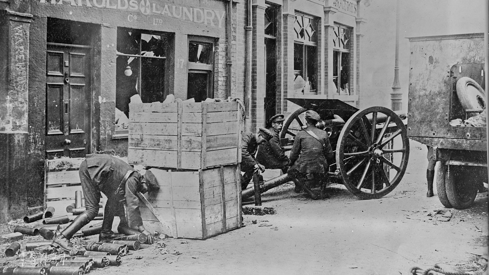 Black and white press photograph of National Army soldiers crouching behind an 18-pounder field gun and wooden crates in a Dublin street, with spent shell cases on the ground.