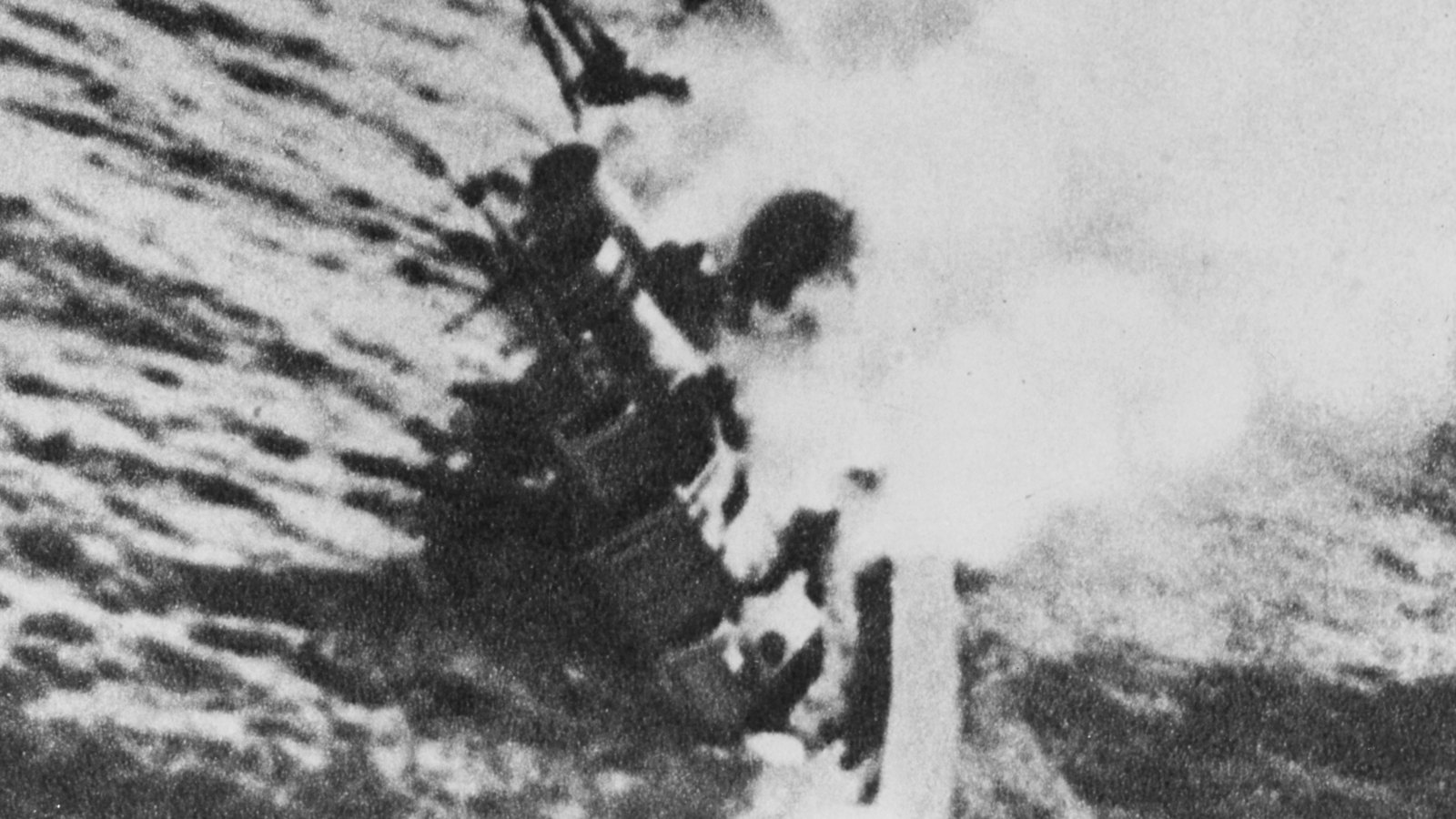 Black and white aerial photograph of a warship listing heavily in a rough sea, her masts tilted and white smoke pouring from her side.