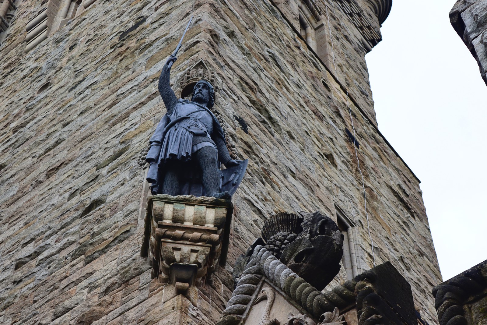 Low-angle photograph of a dark bronze statue of a bearded medieval warrior in mail and a knee-length tunic, raising a sword in his right hand and holding a shield at his side, standing on a carved stone corbel high on the corner of a tall sandstone tower under a pale sky
