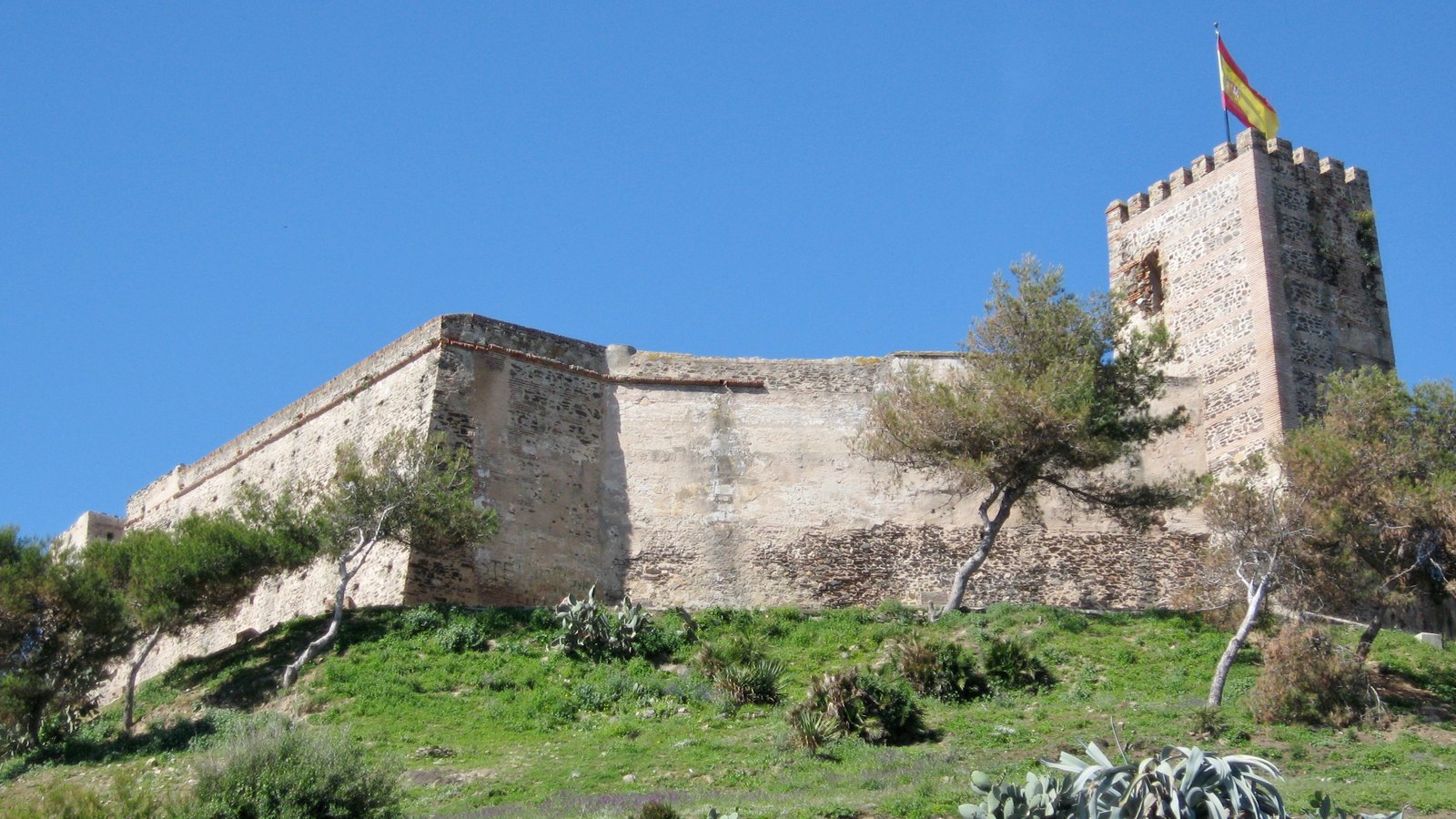 Stone walls and a square crenellated tower of Sohail Castle on a green hillside with agave plants, under a clear blue sky.