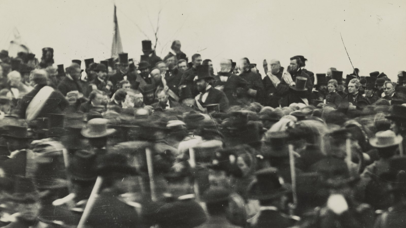 Old photograph of a dense crowd in hats and uniforms in front of a raised platform crowded with men in top hats, a flag on a pole behind them under a pale sky
