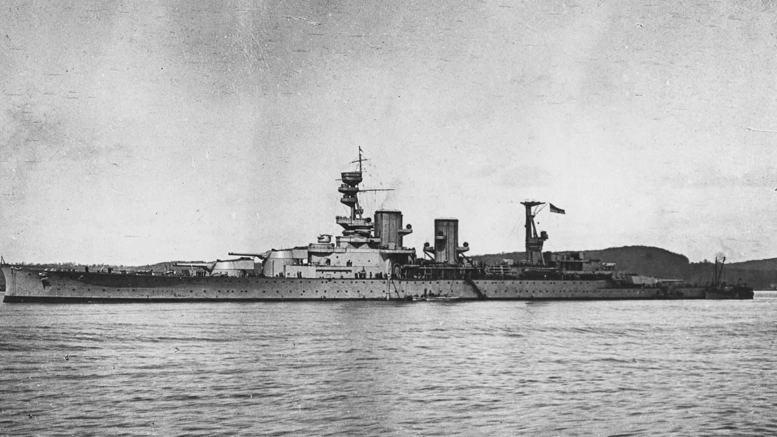Black and white photograph of a long gray battlecruiser with two funnels and a tall tripod mast, lying at anchor on calm water in front of low hills.