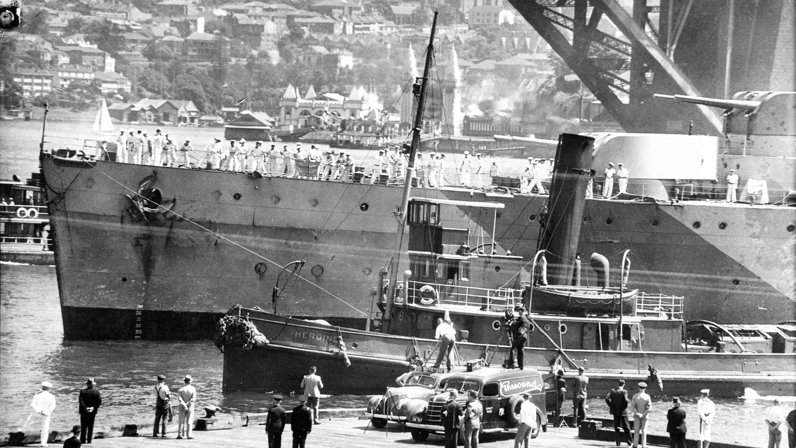 Black and white photograph of a gray cruiser in dazzle camouflage mooring at a Sydney wharf beneath the steel arch of the Harbour Bridge, sailors lining her rail and a crowd and newsreel van on the quay.
