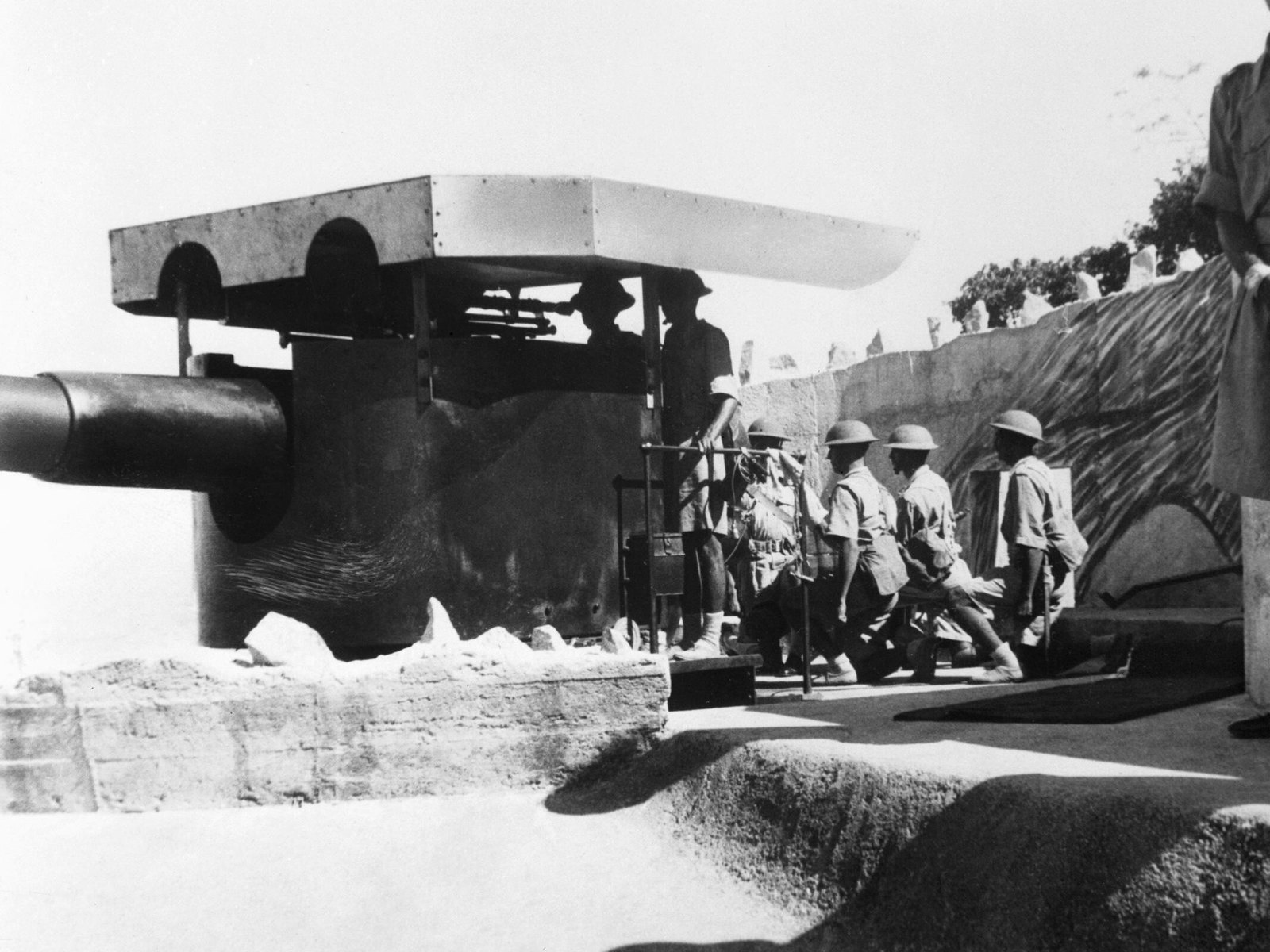 Black and white photograph of soldiers in steel helmets and shorts manning a large coastal gun under a sun shade in a concrete emplacement, with a camouflaged wall behind