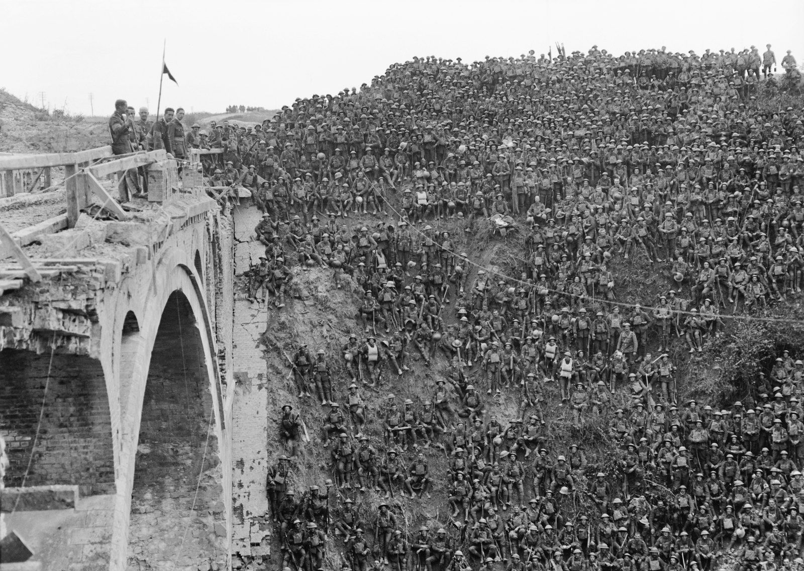 Black and white photograph of hundreds of soldiers in steel helmets crowding the steep bank of a canal cutting beside a damaged brick bridge, where an officer addresses them from the parapet