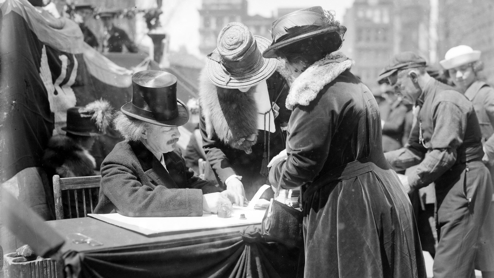 Paderewski w cylindrze siedzi przy przykrytym tkaniną stole na ulicy miasta, a dwie kobiety w kapeluszach i płaszczach z futrzanymi kołnierzami pochylają się nad stołem obok niego