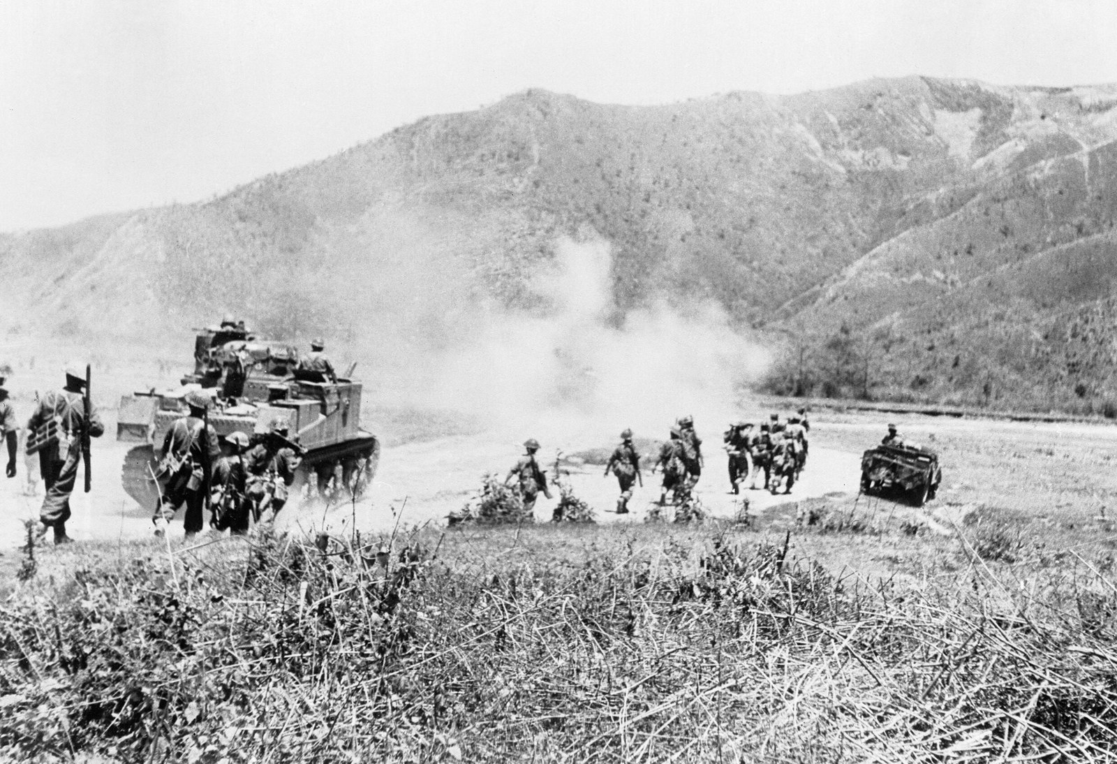 Black and white photograph of infantry advancing across open grassland behind a Lee tank and a carrier, with smoke rising in front of steep wooded hills