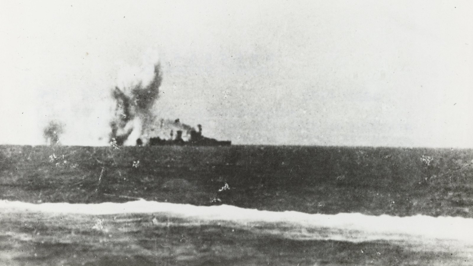 Grainy black and white photograph of a cruiser on the horizon with bomb splashes rising just ahead of her bow, the sea in the foreground.