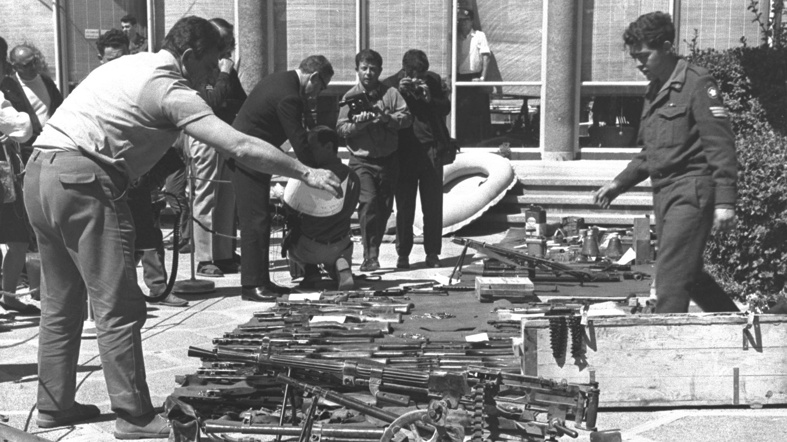 Black and white photograph of a paved courtyard in front of a modern building, with rows of captured mortars, machine guns and rifles laid out on the ground while photographers and onlookers examine them.