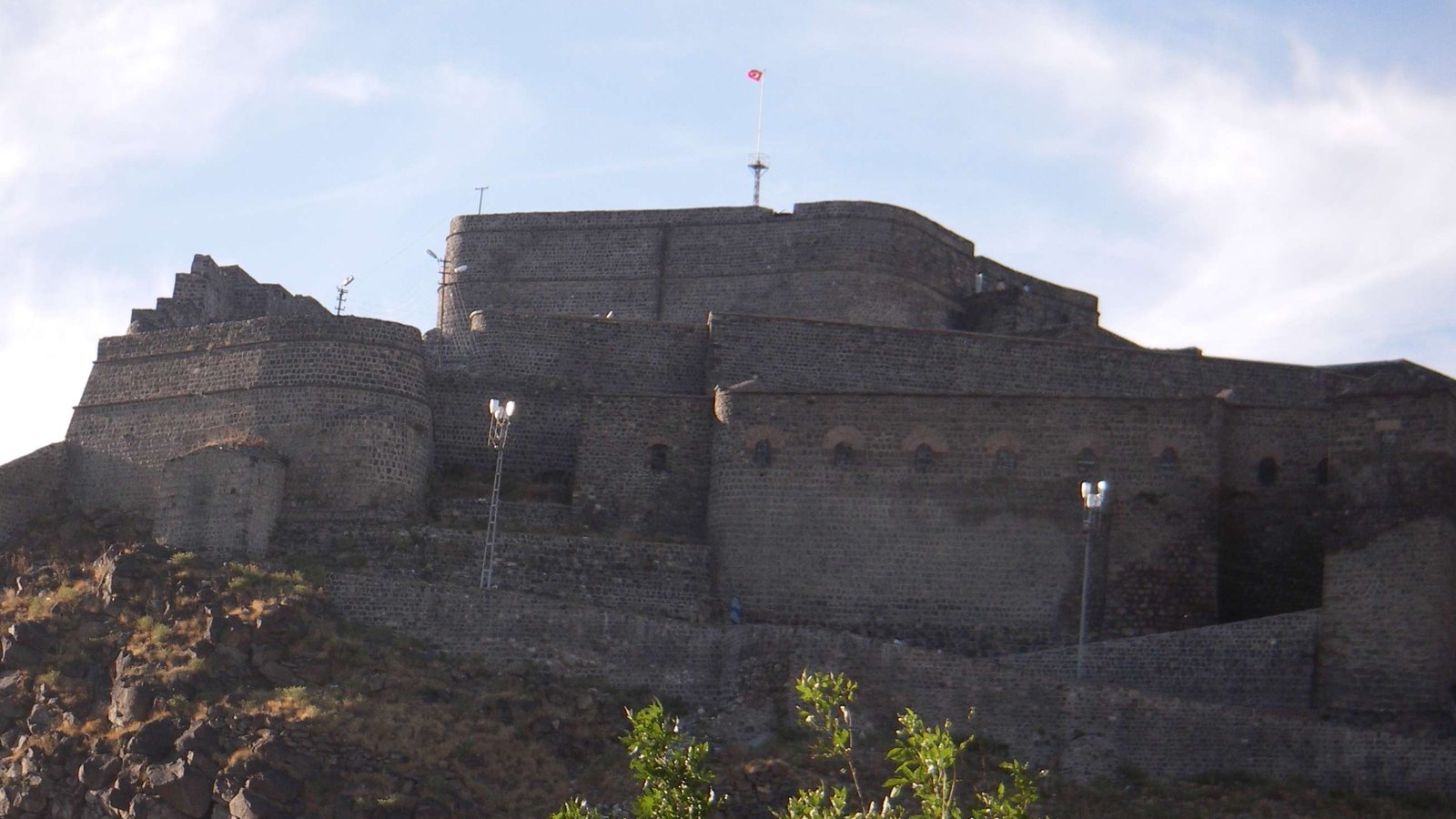Dark basalt walls and towers of the citadel of Kars on a rocky hill under a pale sky, with a red flag on a mast above the keep