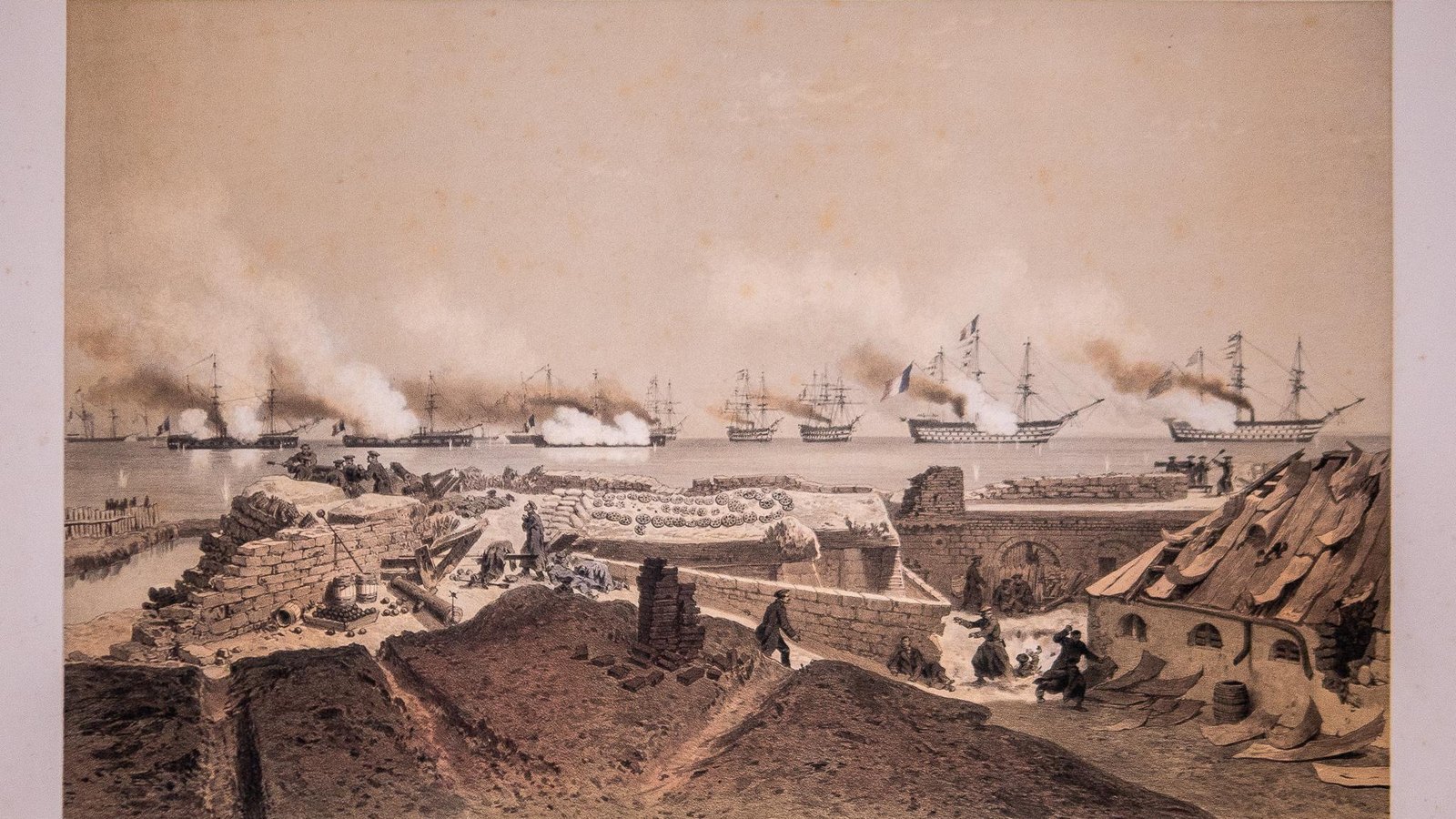Tinted lithograph of a ruined fort with broken walls and a fallen roof in the foreground, soldiers among the ruins, and a line of warships firing in clouds of smoke on the sea behind.