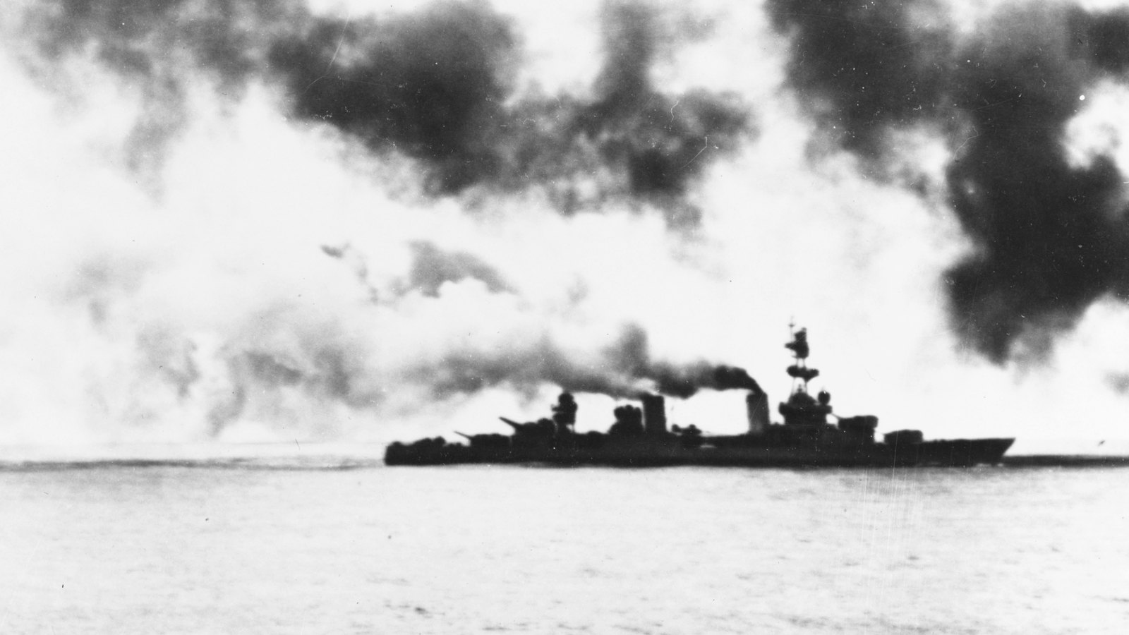 Black and white photograph of the heavy cruiser USS Salt Lake City at sea, silhouetted against heavy smoke, with dark smoke clouds filling the sky above her.