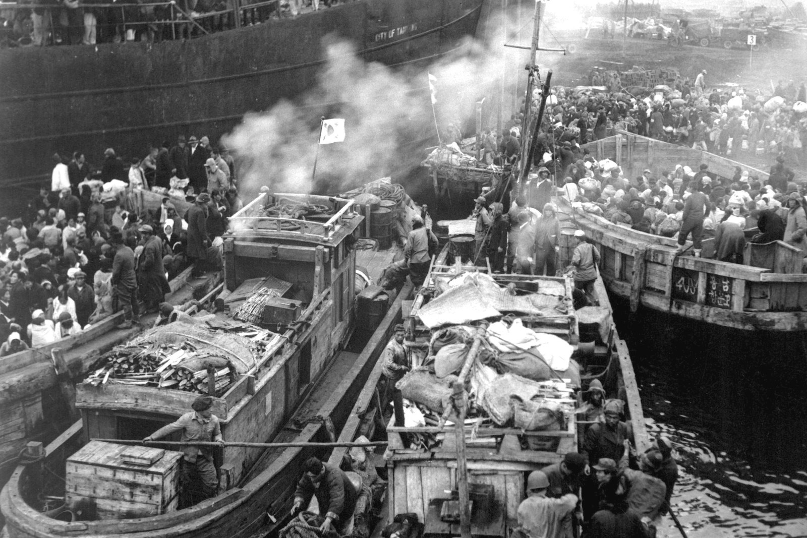 Black and white photograph: crowds of Korean refugees with bundles pack the deck of a large ship and several wooden fishing boats moored alongside it in the harbor of Hungnam, with smoke drifting over the boats.