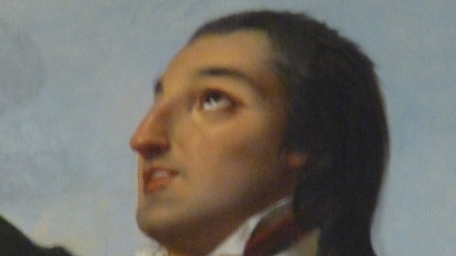Detail of a painted portrait of a young man with long dark hair looking up, wearing a dark coat with a red Sacred Heart badge and a checked scarf, against a pale sky.