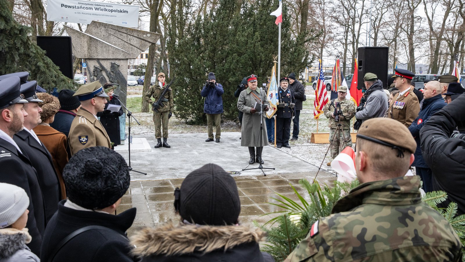 Outdoor ceremony at a stone memorial under a banner reading Powstańcom Wielkopolskim, with a man in a 1919 Polish greatcoat speaking, soldiers, flags and onlookers around him.
