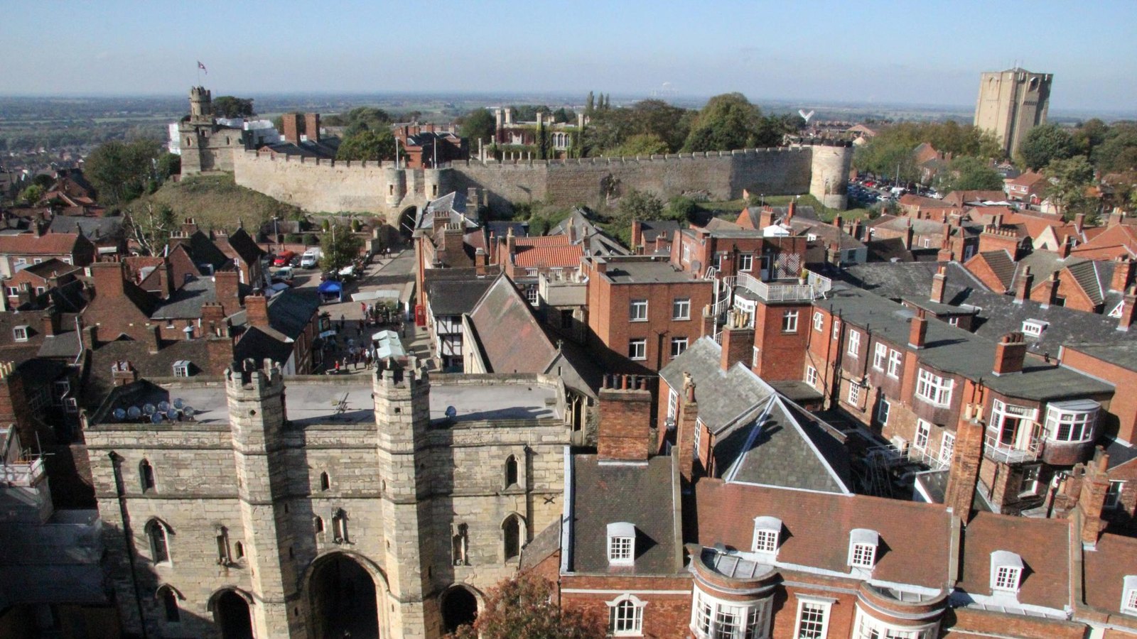 View from a cathedral tower over red brick roofs and a stone gatehouse to the long curtain wall and towers of Lincoln Castle on its hill.