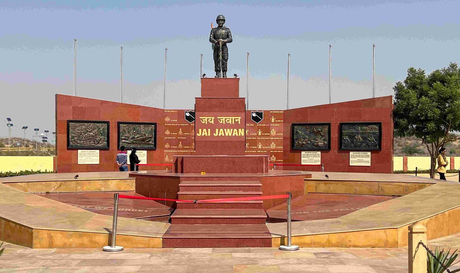 Red stone war memorial at Longewala with a bronze soldier statue on a plinth inscribed Jai Jawan, bronze relief panels and a row of flagpoles under a pale sky