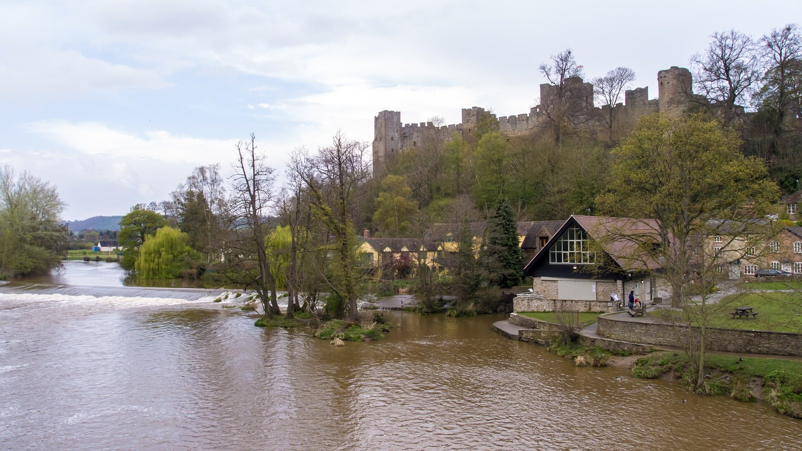 Ruined battlements of Ludlow Castle above trees and riverside houses, with a brown, swollen river and a weir in the foreground under a cloudy sky.