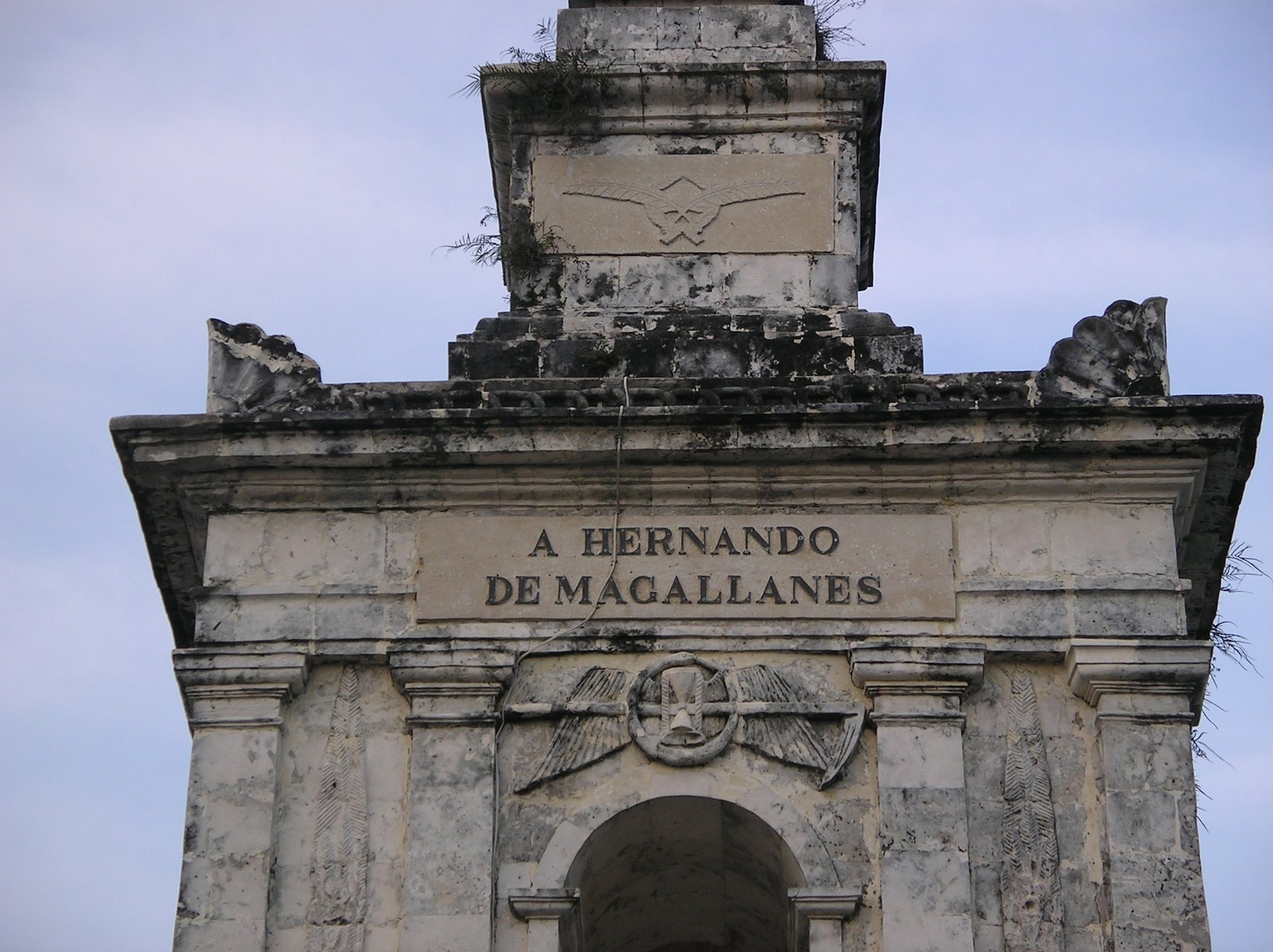 Weathered gray stone monument with an arched base, carved winged emblems and an inscription reading A Hernando de Magallanes, against a pale blue sky