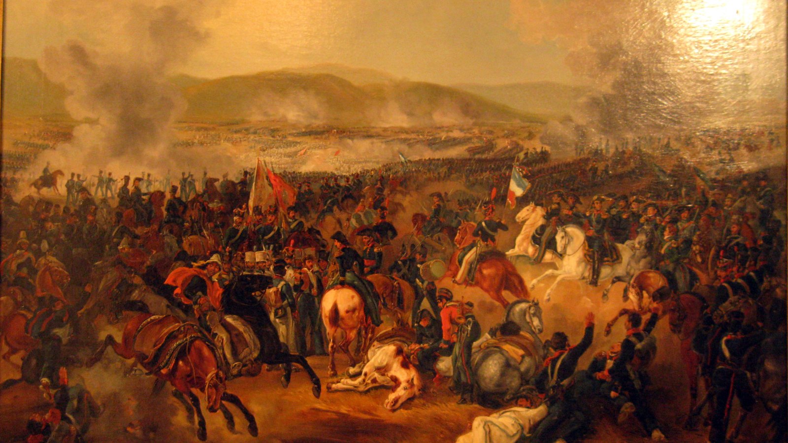 Nineteenth-century oil painting of the Battle of Maipú: a dense crowd of cavalry and infantry with flags in the foreground, fallen horses and men, and smoke rising over a valley and hills behind.