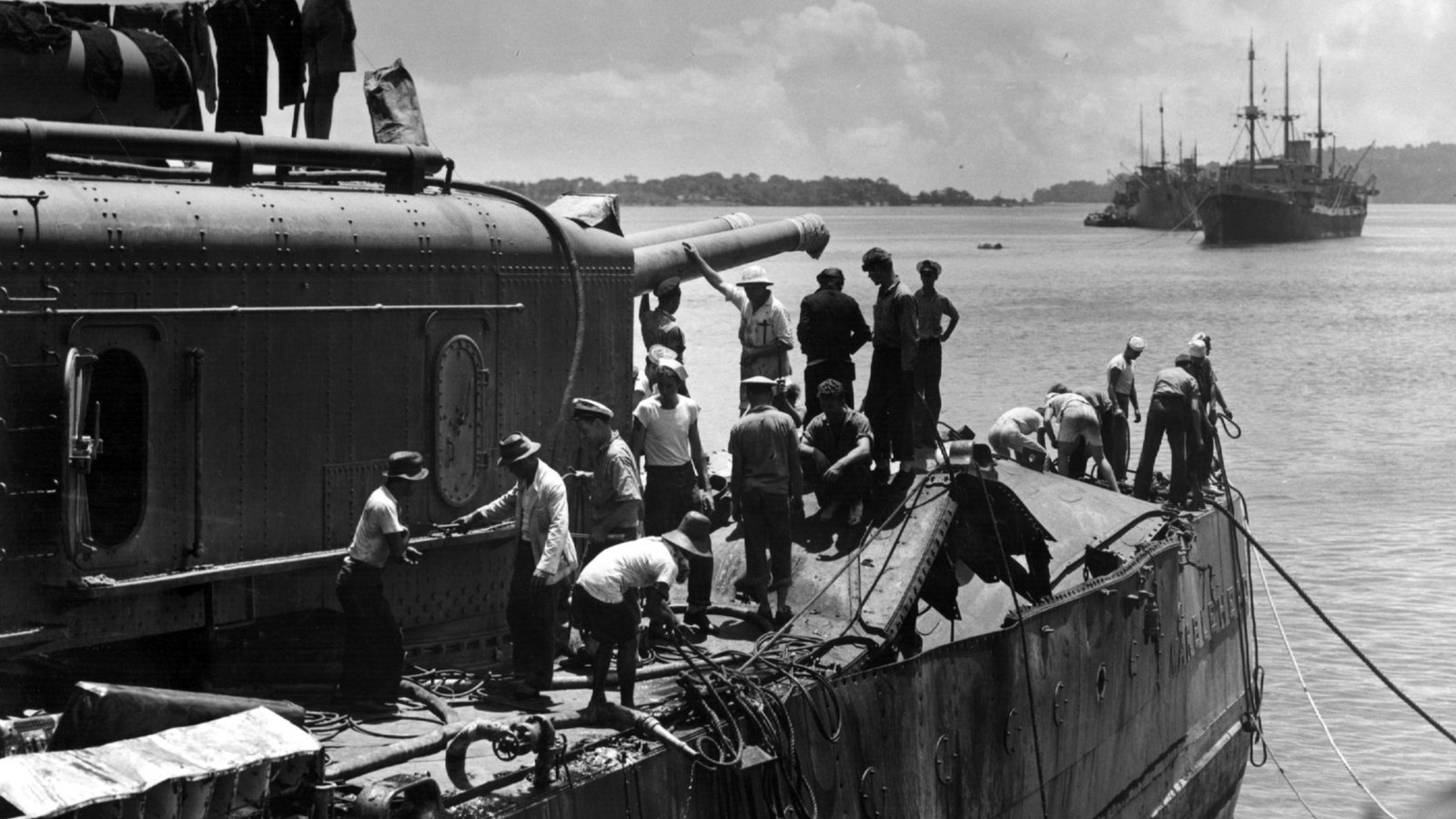 Black and white photograph of the stern of a damaged warship moored in a harbor, a gun turret on the left and men in shirtsleeves and helmets working on torn deck plating, two cargo ships at anchor behind.