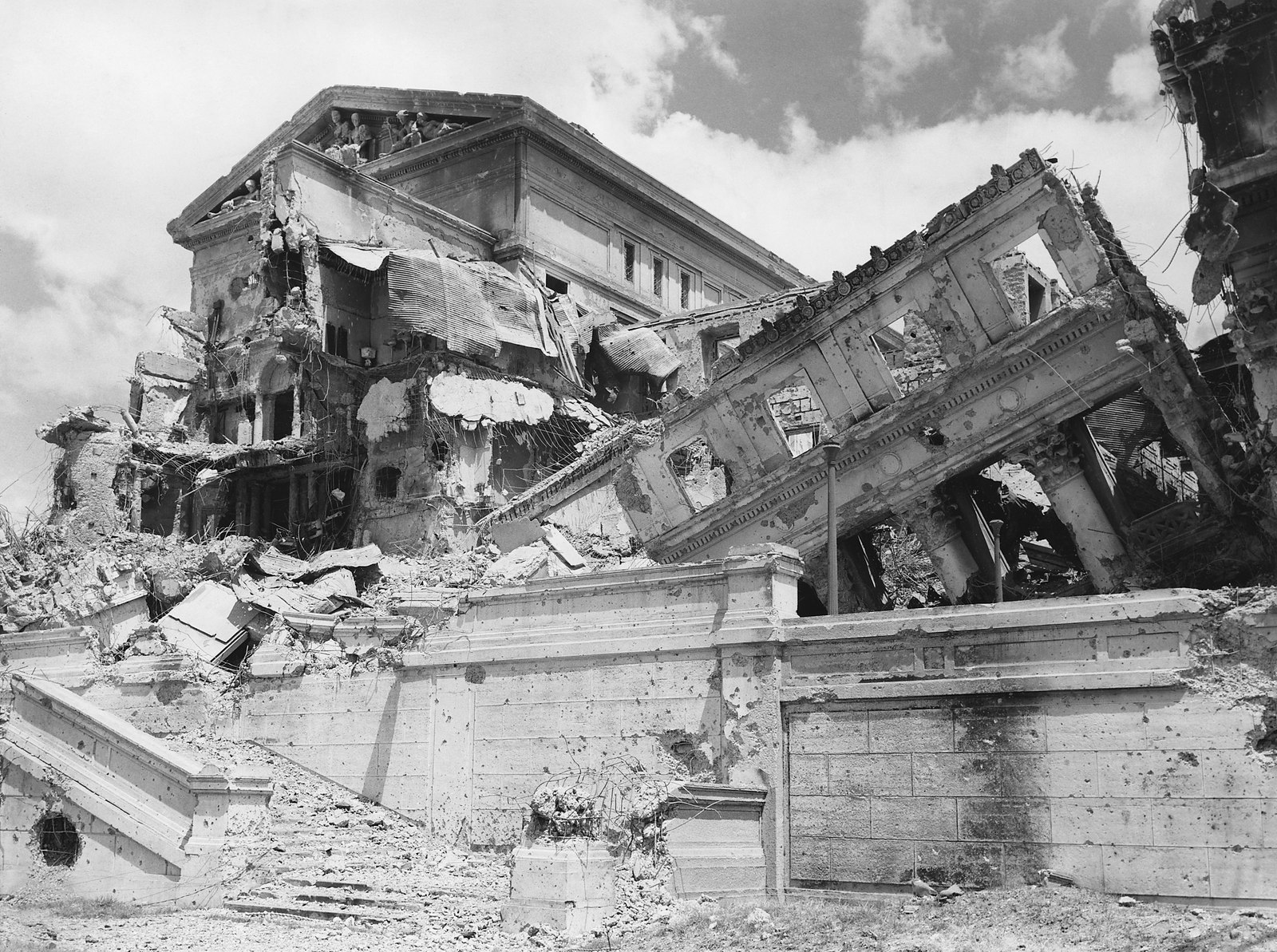 Black and white photograph of the shattered Legislative Building in Manila: a collapsed facade leans over a pockmarked stone terrace, with broken concrete heaped below and clouds overhead