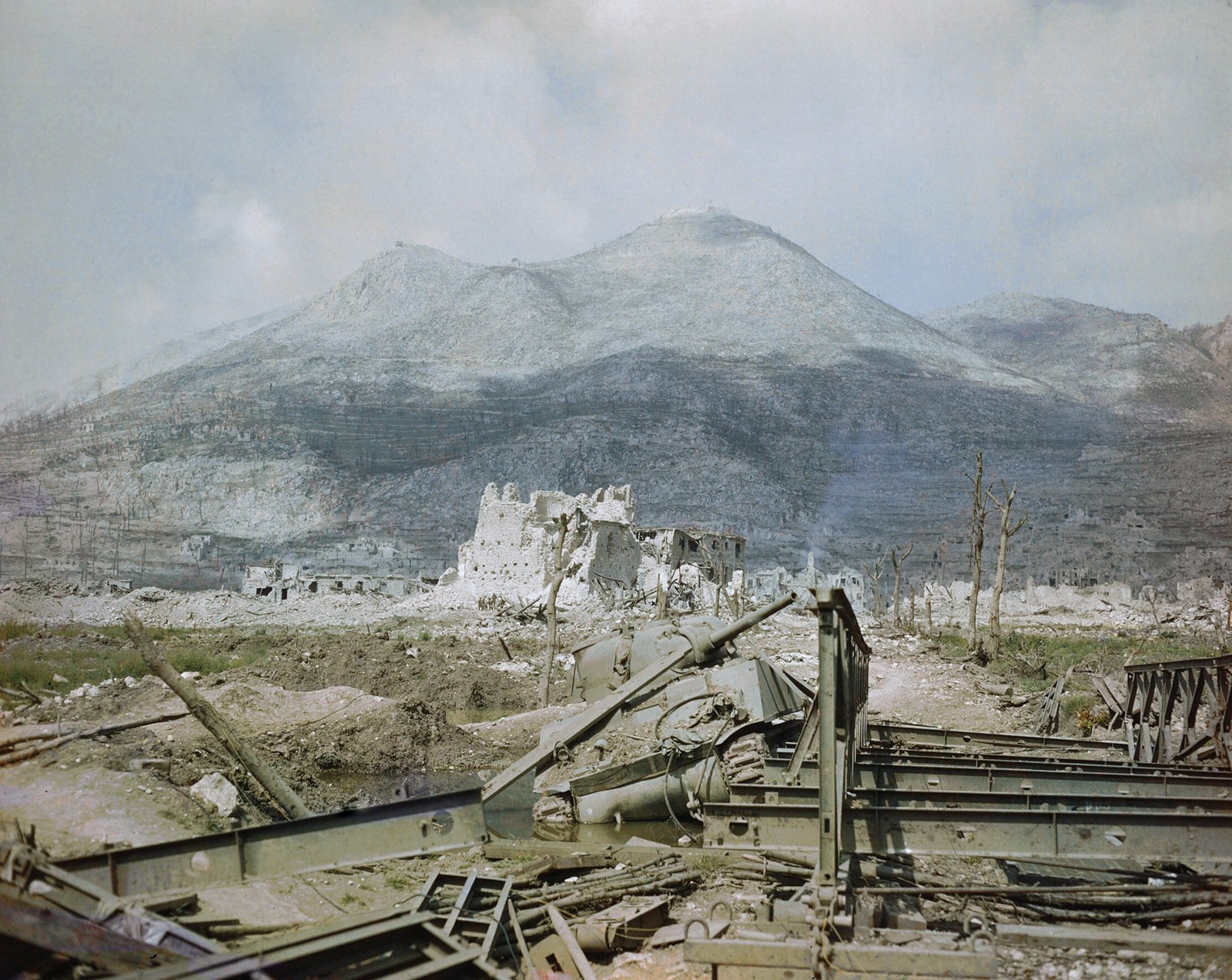 Barwne zdjęcie zrujnowanego Cassino w maju 1944 roku: na pierwszym planie zniszczony czołg Sherman i powyginane przęsła mostu Bailey, za nimi zburzone domy i nagie drzewa, a nad miastem szare zbocza Wzgórza Klasztornego.