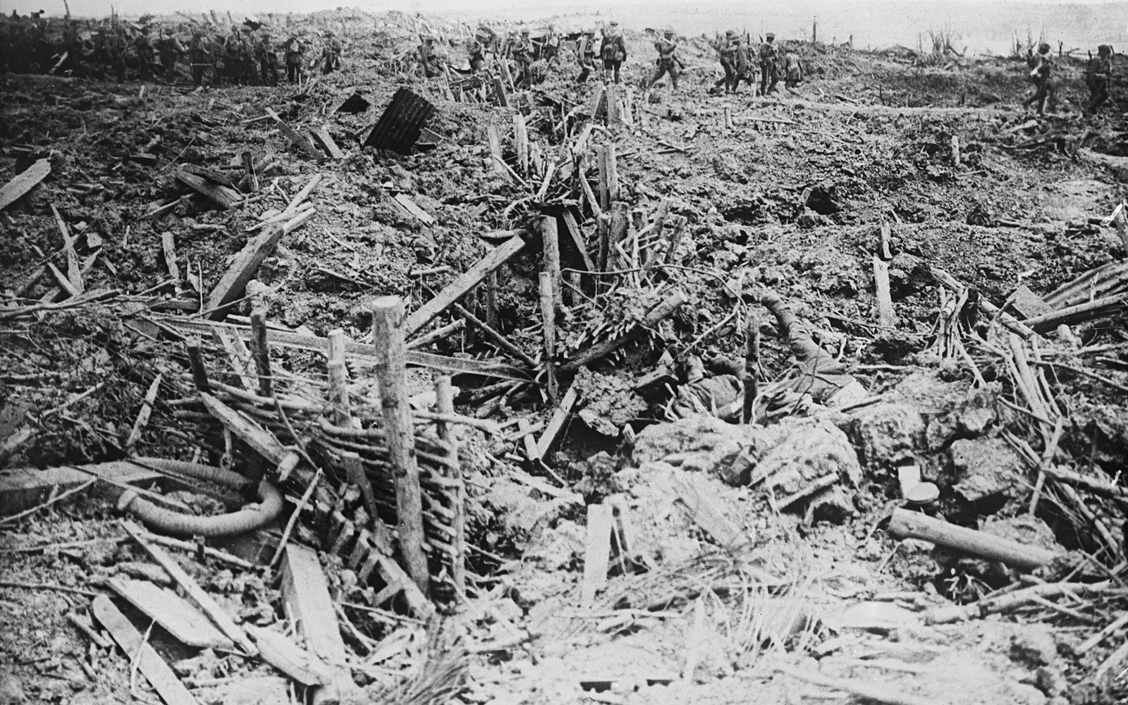 Black and white photograph of British infantry in steel helmets walking in file across the crest of a ridge of churned earth, with smashed trench timbers, wicker revetting and broken stakes in the foreground