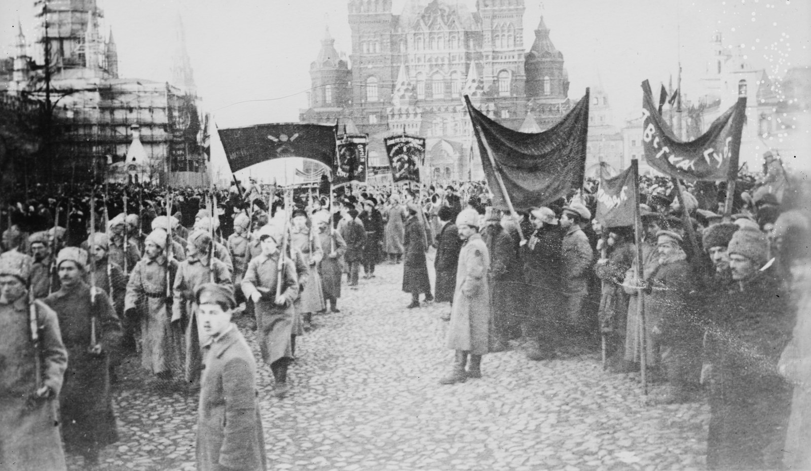 Czarno-białe zdjęcie żołnierzy w długich płaszczach i futrzanych czapkach maszerujących z karabinami przez brukowany plac obok tłumu z ciemnymi sztandarami, w tle wieże Państwowego Muzeum Historycznego w Moskwie.