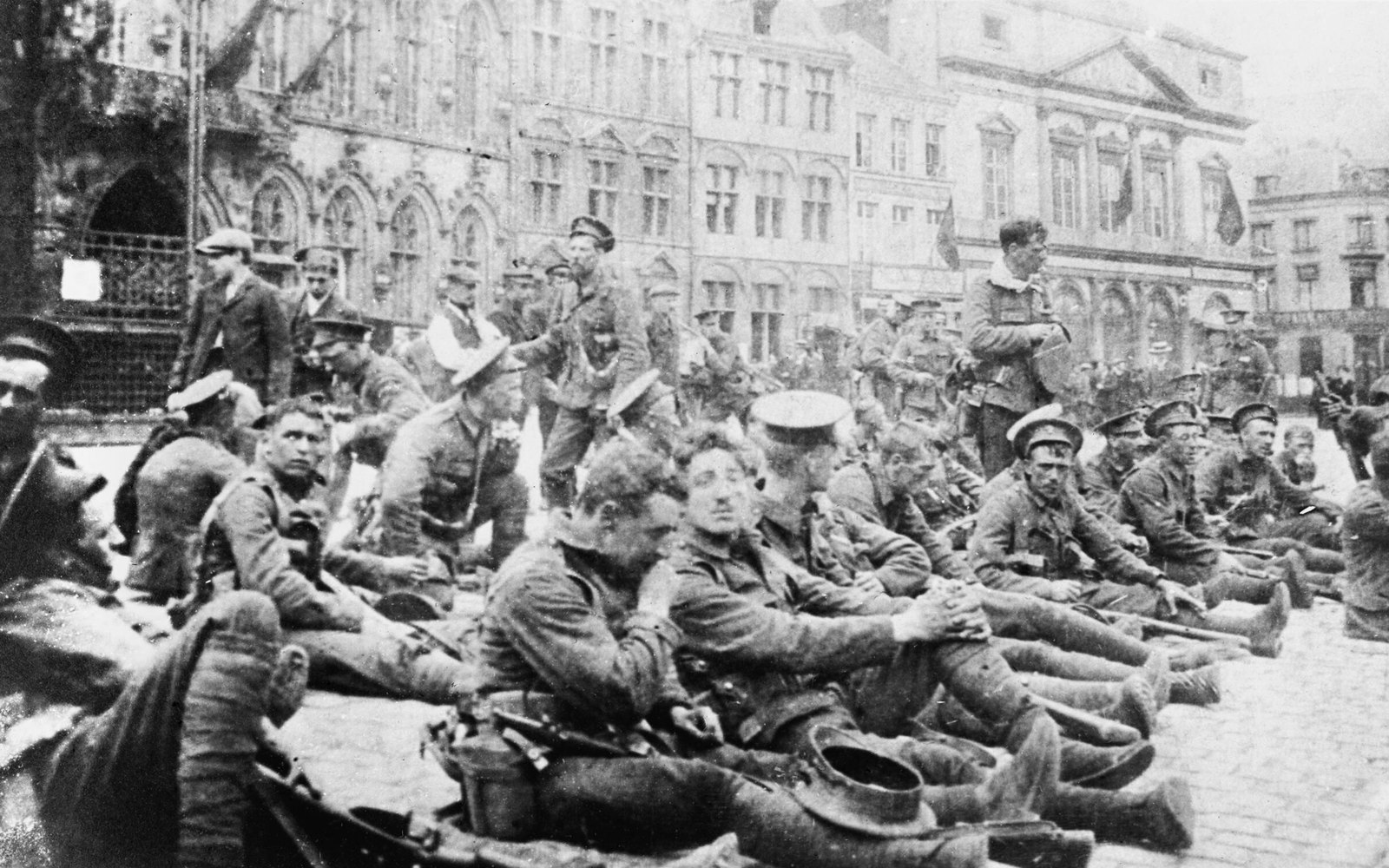 Czarno-białe zdjęcie brytyjskich żołnierzy w czapkach z daszkiem i mundurach khaki, siedzących i odpoczywających na bruku miejskiego placu, z karabinami i oporządzeniem obok, na tle wysokich gotyckich i klasycystycznych kamienic z flagami