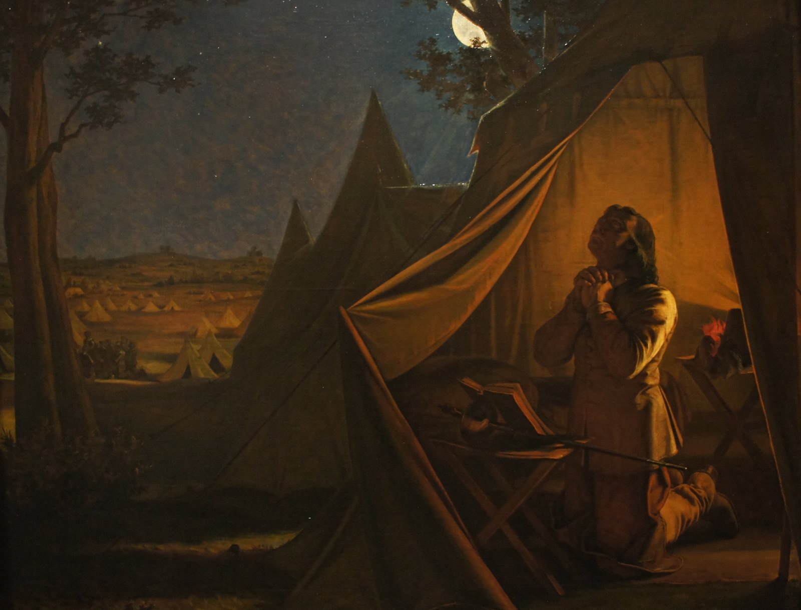 Oil painting of a night camp: a long-haired man in a buff coat kneels with clasped hands and upturned face in the warm light of an open tent, a sword and an open book on a folding stool beside him and a plumed helmet behind, while rows of pale tents stretch across the fields under a starry sky and a moon half hidden by a tree