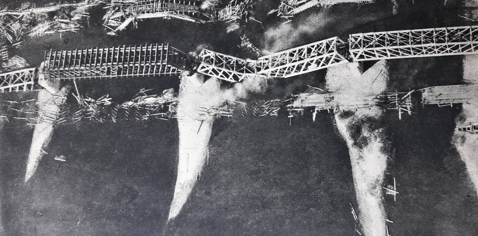 Aerial photograph of a river with a collapsed iron truss bridge on stone piers, burned wooden bridges and floating debris